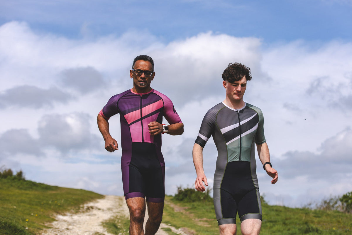 Two triathletes wearing SLS3 tri suits jogging outdoors along a grassy coastal trail under a partly cloudy sky.