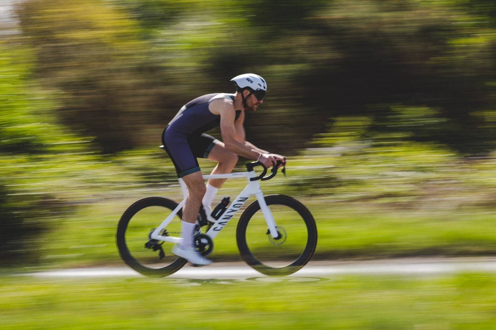 A male triathlete in a dark SLS3 tri suit and white helmet cycling at speed on a white road bike, with motion-blurred greenery in the background.